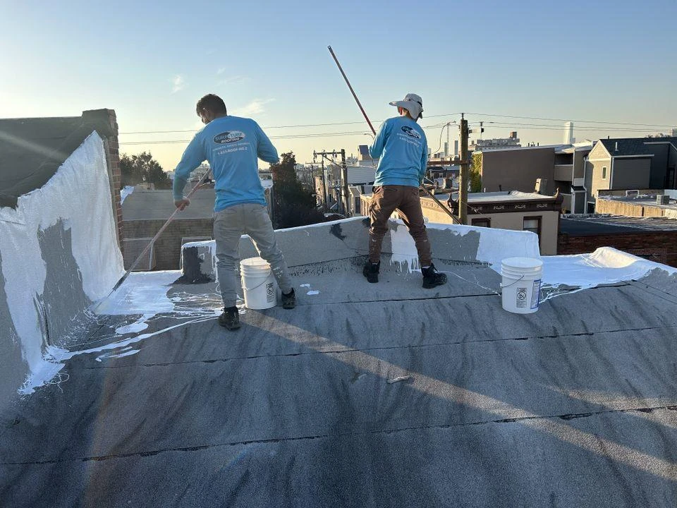 Two roofing professionals in blue shirts working on flat roofs repairs, applying a white protective coating under clear skies in a residential area.