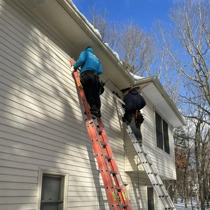 Two roofing contractors on ladders replacing a roof in winter conditions, showing that 'can you replace roof in winter' is feasible with the right expertise and safety measures.